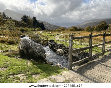 A countryside scene with an old wooden bridge over a small river and mountains in the distance.  Royalty-Free Stock Photo #2274651697