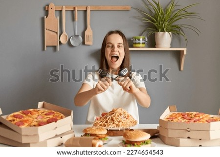Portrait of amazed overjoyed woman with brown hair wearing white casual T-shirt sitting at table in kitchen, holding to forks, crossed hands, yelling with crazy expression, being extremely hungry.