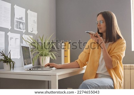 Adorable brown haired businesswoman holding smartphone, recording voice message in social network, chatting online by speakerphone, woman sitting at desk with laptop in office.
