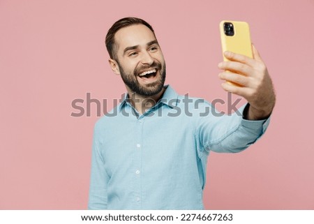 Young smiling cheerful fun caucasian man 20s wear classic blue shirt doing selfie shot on mobile cell phone post photo on social network isolated on plain pastel light pink background studio portrait.