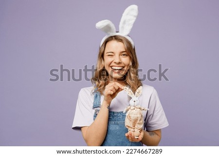 Young smiling cheerful fun woman wear casual clothes bunny rabbit ears hold in hands toy wink blink eye isolated on plain pastel light purple background studio portrait. Lifestyle Happy Easter concept