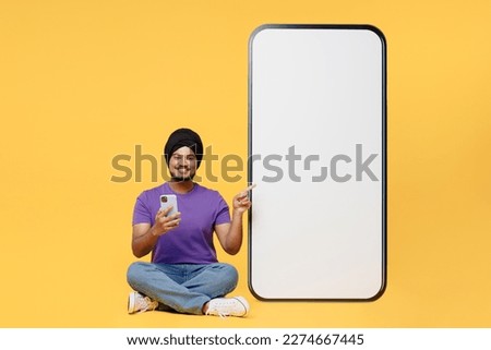 Full body devotee Sikh Indian man ties his traditional turban dastar wear purple t-shirt show big huge blank screen mobile cell phone use smartohone isolated on plain yellow background studio portrait Royalty-Free Stock Photo #2274667445