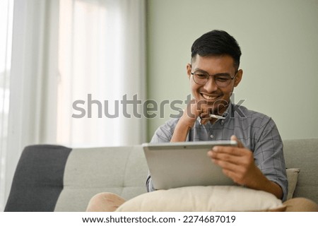 Happy and smiling millennial Asian man in casual clothes using his digital tablet, watching video or working on his project, sitting on sofa in his living room.