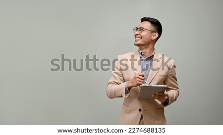 Smiling and handsome millennial Asian businessman in a formal business suit and glasses, looking aside at the empty space, holding a tablet, stands against a green studio background. Royalty-Free Stock Photo #2274688135