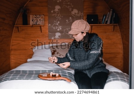caucasian girl in woolen sweater and brown hat sitting on cozy log cabin bed while spreading cheese and barbecue sauce on appetizing crispy small thin toast before having a snack, te wepu pods akaroa
