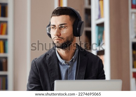 Bearded man with eyeglasses sitting in library, working on laptop and listening to audio course or podcast with headphones, making pause and looking away. Student studying, preparing assignment