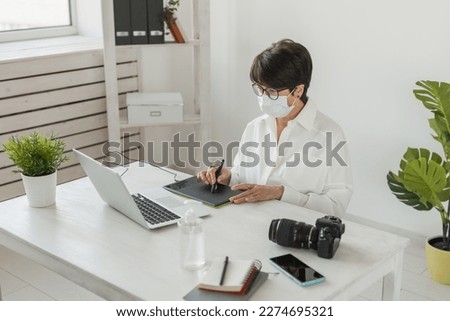 Mature intelligent woman with medical mask working with computer and graphic tablet and stylus. She is a successful self employed retoucher and photograph