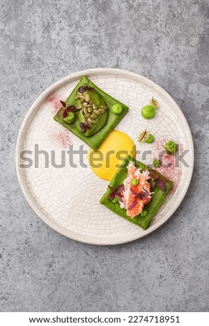 Colorful green crepes or blini with crab meat, avocado mousse and hollandaise sauce, decorated with micro greens and pumpkin seeds over gray concrete background. Gourmet snack. Fine dining. Top view Royalty-Free Stock Photo #2274718951