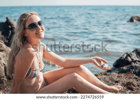 A blonde girl in a striped swimsuit and glasses sits on the ocean and smiles. Pebbles and waves all around on a bright sunny day.