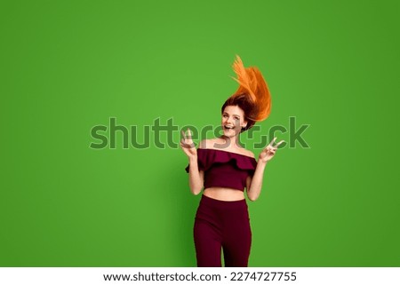 Red straight-haired attractive cute nice cheerful young girl, half turned, wind blowing hair up in air, showing double v-sign. Isolated over bright vivid yellow background
