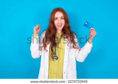 Photo of lucky impressed young doctor woman wearing medical uniform over blue background arm fist holding credit card. Celebrated