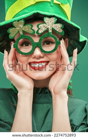 Close-up portrait of a funny pretty girl dressed in a green leprechaun hat and green clover-shaped glasses smiles at the camera. Studio portrait on a green background. Party for St. Patrick's Day.