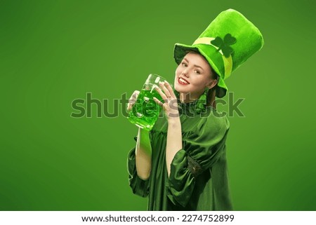 Cheerful attractive girl dressed in a green dress and a leprechaun hat holds a beer and smiles at the camera. Party for St. Patrick's Day. Studio portrait on a green background with copy space. 