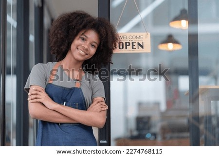 Portrait of African American businesswoman standing at the cafeteria door entrance. A cheerful young waitress in a blue apron near a glass door with an open signboard and looking at the camera. Royalty-Free Stock Photo #2274768115