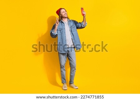 Photo of cheerful satisfied optimistic guy long hairstyle jeans shirt making selfie on front camera isolated on yellow color background