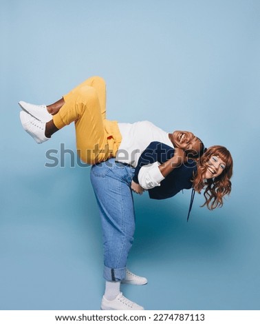 Playful young woman carrying her best friend on her back in a studio. Two vibrant young women having fun against a studio background. Female best friends making happy memories together. Royalty-Free Stock Photo #2274787113