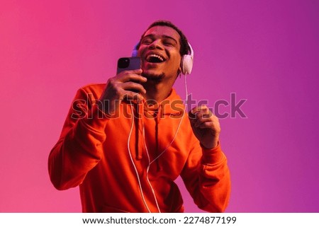 Young african man listening music with headphones and singing while holding smartphone like microphone isolated over blue neon wall