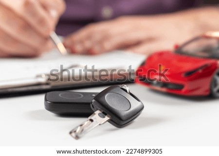Car keys on desk with man signing purchase documents in background. Closeup of black modern car keys while hand complete the insurance policy or rental documents. Guy buying new car at dealership.