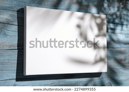 Canvas mockup, white blank picture hanging on blue wooden wall with dark shadows of palm leaves. Poster mock up, empty canvas with shadows of plant