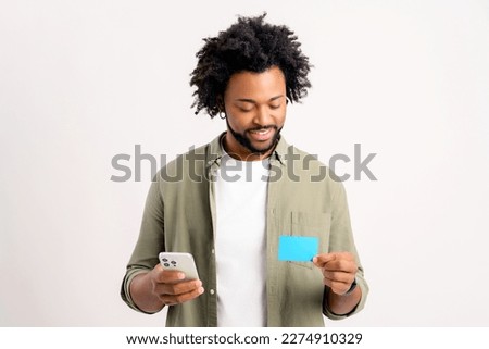 Blissful young multiracial man holding debit card and smartphone isolated. Lucky man in casual clothes receive cash back from online purchases Royalty-Free Stock Photo #2274910329
