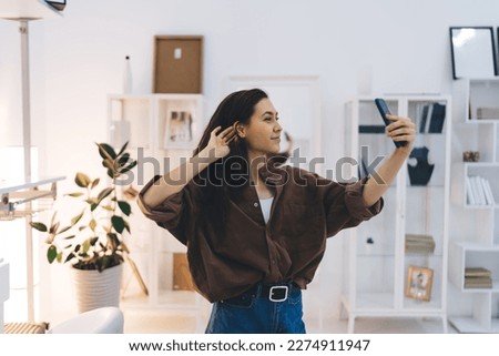 Smiling stylish young brunette in trendy clothes standing in contemporary light room and taking selfie on smartphone while enjoying weekend at home