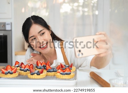 Cheerful young girl wearing apron taking a selfie and showing baked fruit tart in white bright kitchen, holding mobile phone taking a selfie, .Smiling sian woman bakery shop owner making bakery.