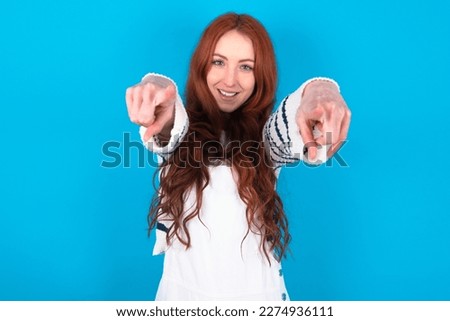 Close-up portrait of surprised young caucasian woman wearing overalls over blue background pointing with two fingers to the camera saying: I choose you!, looking up with open mouth.