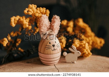 Close-up horizontal photo of wooden Easter bunny with pink ears. It stands on a wooden board together with small white wooden rabbit. Background is Mimosa spring flowers