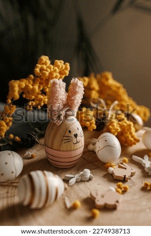 Vertical photo of wooden Easter bunny with pink ears. It stands on a wooden stand and  surrounded by small white and gold Easter eggs. Background is yellow Mimosa spring flowers