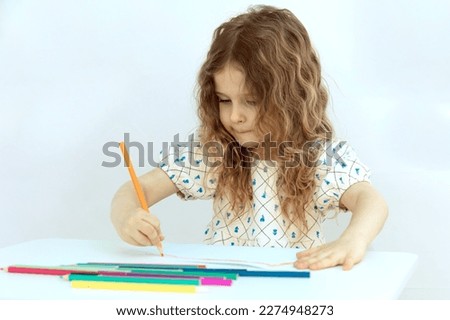 Blonde child girl drawings with colored pencils sitting at the table on a white background. Concept of preparation for school . Children education