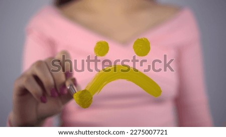 A young woman draws a sad smile on the glass. A girl in a pink sweater draws with a brush and yellow paint. Close-up.