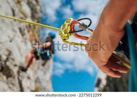 Belay device close-up shot with a boy on the cliff climbing wall. He hanging on a rope in a climbing harness and his partner belaying him on the ground. Active people and sports concept image Royalty-Free Stock Photo #2275012299