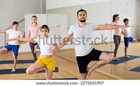 Young girl holding hand of her father while standing in tree pose during family yoga training in fitness studio.