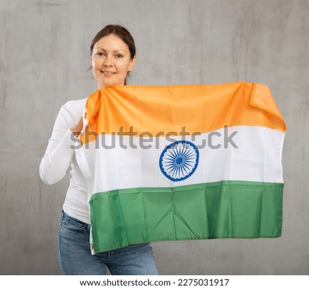 Happy young woman with large flag of India posing gladly against light unicoloured background