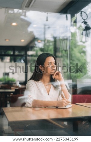Vertical portrait of an attractive female writer looking for inspiration to start a new novel on her notepad. Young woman meditating in a coffee shop