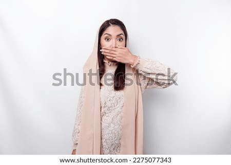 Oops! Surprised young Asian Muslim woman covering mouth with hands and staring at camera while standing against white background