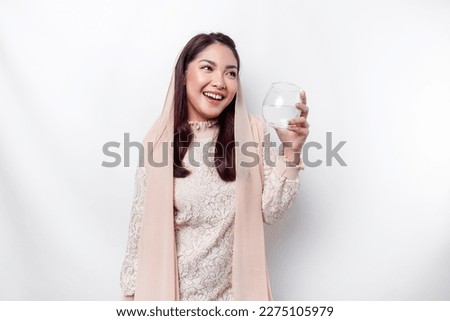 Joyful Asian Muslim woman wearing headscarf is drinking a glass of water, isolated on white background. 