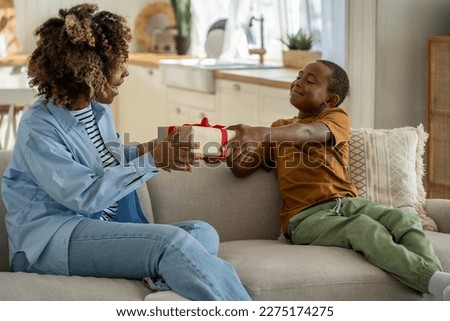 For the best mom ever. Proud happy African American boy son giving wrapped gift box to surprised excited mother, sitting together on couch at home. Child kid surprising mum on Mothers day