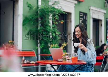 Happy young caucasian woman relaxing in outdoor cafe and drinking coffee while working on a laptop. Portrait of a businesswoman working outside during a sunny day. Copy space.