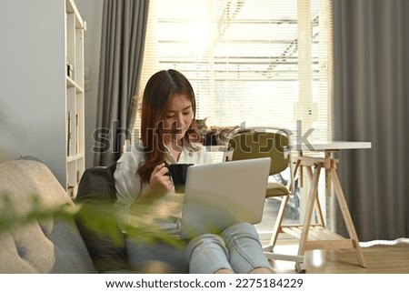Young asian woman freelancer sitting on couch surfing internet, checking email on laptop in the morning