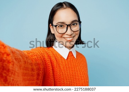 Close up young fun woman of Asian ethnicity wear orange sweater glasses doing selfie shot pov on mobile cell phone isolated on plain pastel light blue cyan background studio. People lifestyle concept