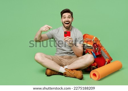 Full body young traveler white man near backpack stuff mat sit point finger on mobile cell phone isolated on plain green background Tourist lead active lifestyle Hiking trek rest travel trip concept Royalty-Free Stock Photo #2275188209