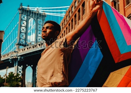 Young black man with arms outstretched holding Progress Pride flag with Brooklyn Bridge in background Royalty-Free Stock Photo #2275223767