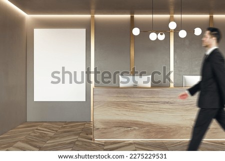 Businessman walking by stylish natural wooden reception desk with modern computer in office with blank white poster with place for your logo or text on beige wall background with golden decoration