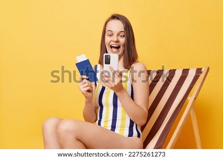 Image of amazed happy young woman wearing striped one-piece swimsuit, holding passport and using cell phone, booking tickets for traveling, sitting on wooden chair isolated on yellow background