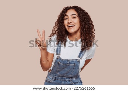 Portrait of young happy smiling woman with curly hair, does peace victory sign with two fingers, at studio isolated over beige background.