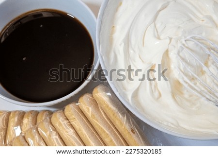 close up of tiramisu ingredients: a cup of coffee, pack of savoiardi biscuits and mascarpone soft cream cheese in bowl, cooking process in the kitchen  