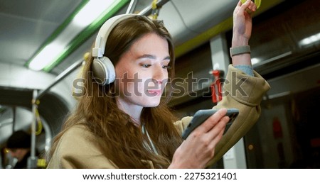 Side-view portrait of focused cute woman watching video on smartphone using headphones riding bus. Attractive happy caucasian female standing in public transport holding handrail. Royalty-Free Stock Photo #2275321401