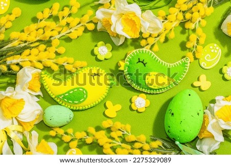 Easter composition with traditional spring flowers and handmade felt birds. Decorative eggs, rabbits, hard light, dark shadow, green background, top view