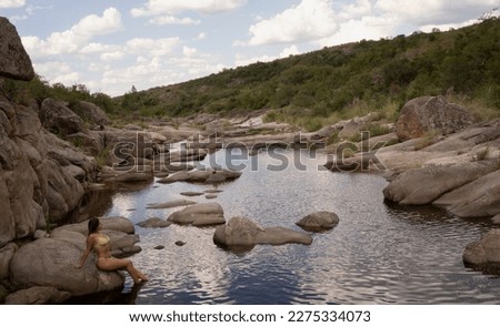 Portrait of a young woman in her swimsuit, relaxing in the river flowing across the rocky hills and cliffs. Beautiful sky reflection in the water surface. 